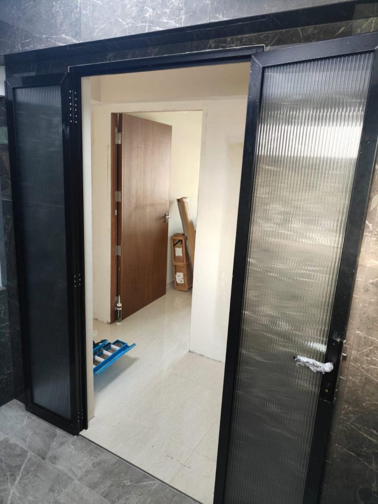 An open set of newly installed Black Aluminium-Framed Fluted Glass Doors in a residential hallway, featuring dark frames and textured glass that reveals an adjacent room undergoing renovation.