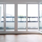 a room with large, white aluminium windows and a glass door, which opens onto a balcony or terrace. The floor is made of wood, and the view through the windows is of other buildings and greenery.