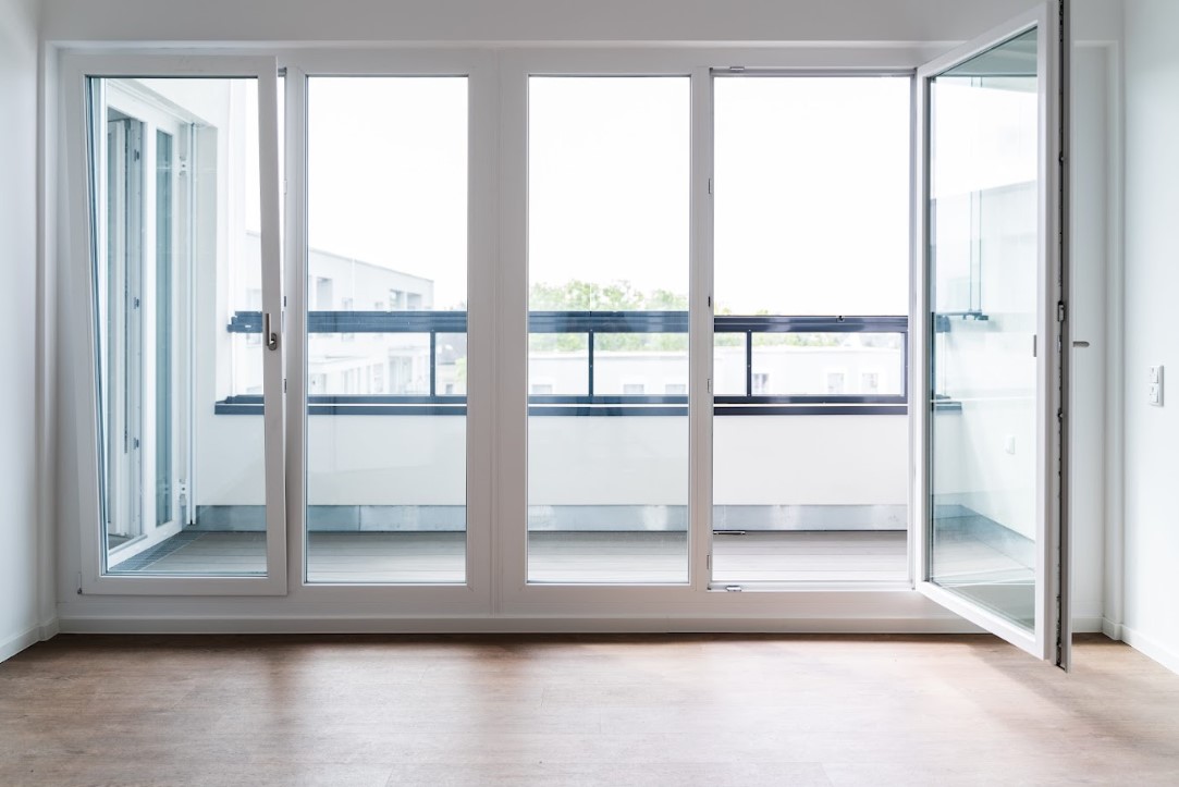 a room with large, white aluminium windows and a glass door, which opens onto a balcony or terrace. The floor is made of wood, and the view through the windows is of other buildings and greenery.
