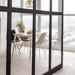 Modern interior with a white dining table and chairs, viewed through black aluminium-framed glass partition doors.
