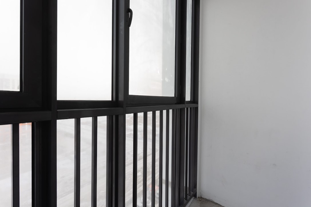 A close-up view of a modern black aluminium window frame and a matching black metal balcony railing against a plain white wall.