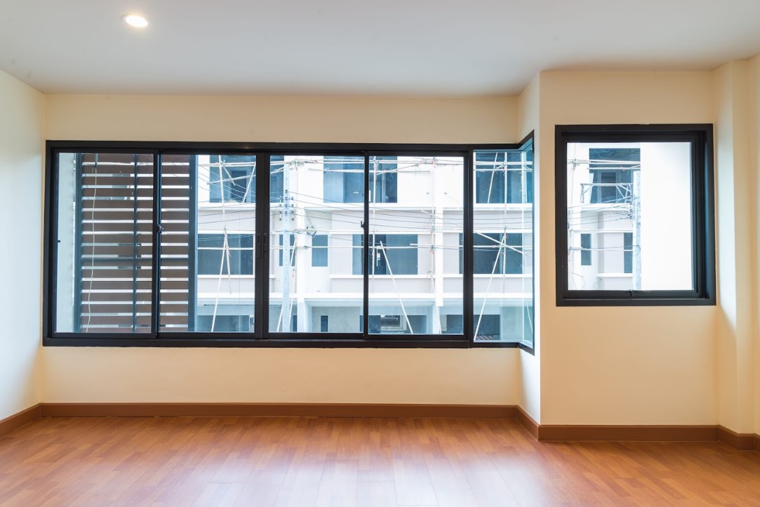 An empty room with wood laminate flooring and cream-colored walls, featuring two large black-framed windows with aluminium grilles