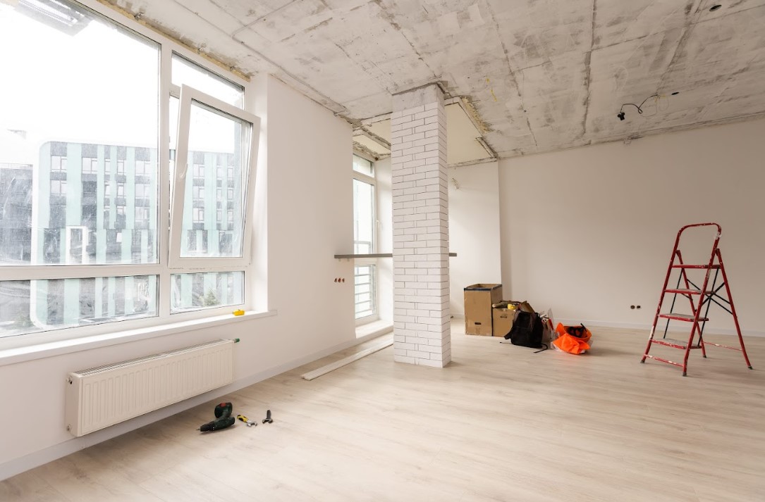 Home renovation in progress inside an empty apartment, showing newly installed windows, a ladder, tools, and construction materials in a bright unfinished interior space.