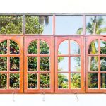 Wooden arched window panels with glass panes reflecting lush tropical greenery and palm trees, showcasing a traditional design suitable for window replacement inspiration.