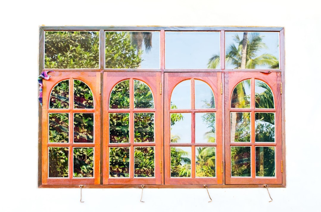 Wooden arched window panels with glass panes reflecting lush tropical greenery and palm trees, showcasing a traditional design suitable for window replacement inspiration.